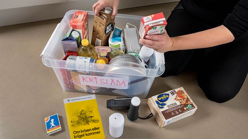 En plastlåda märkt “KRISLÅDA” fylld med matvaror, ljus och ficklampa. Utanför lådan ligger en broschyr “Om krisen eller kriget kommer”, tändstickor och knäckebröd. En person packar ner varor i lådan.