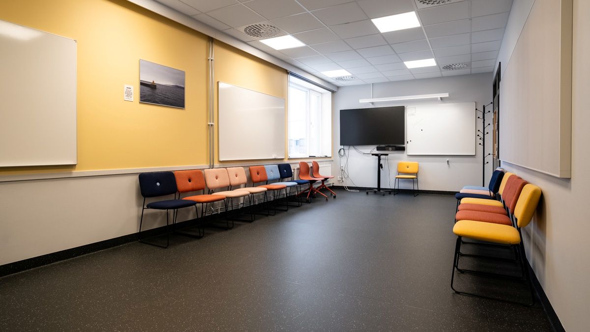 Ett möblerat mötesrum med färgglada stolar placerade längs väggarna, flera whiteboards och en stor skärm vid fönstret.