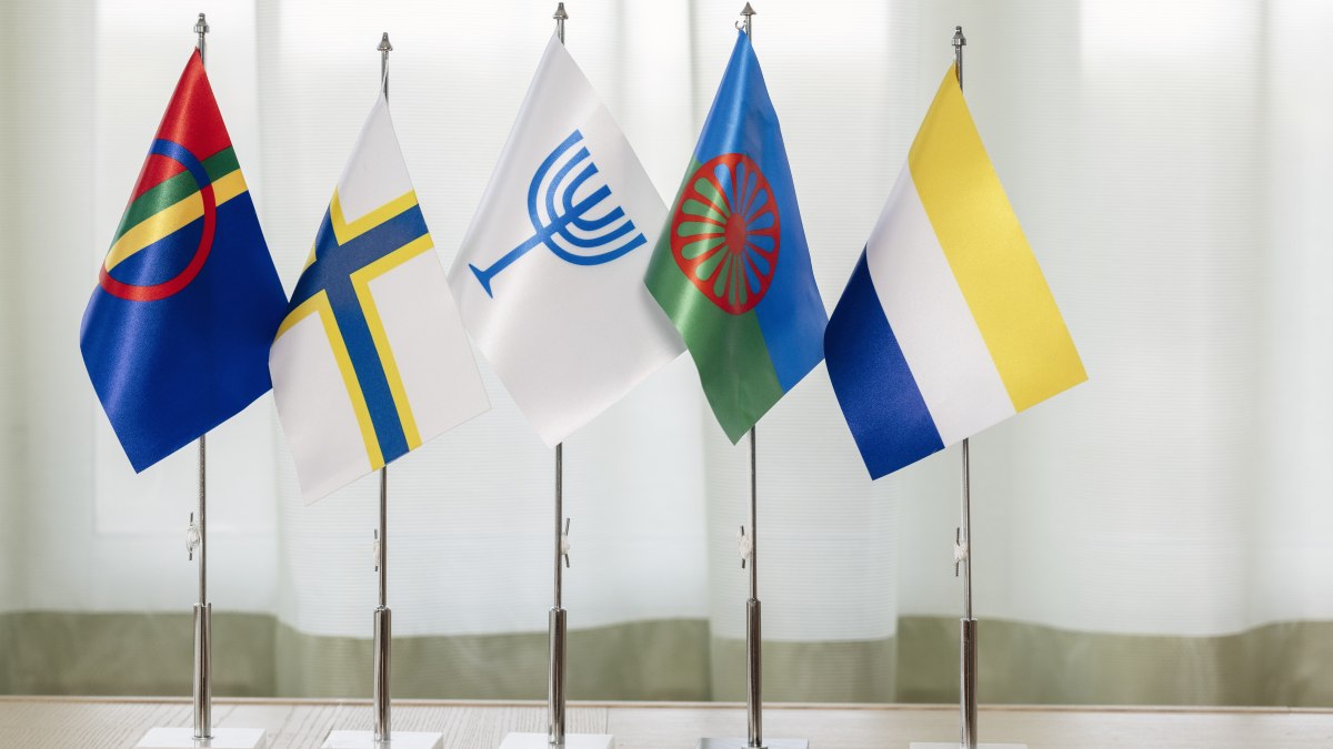 Bordsflaggor med symboler för samer, sverigefinnar, judar, romer och tornedalingar står på ett bord framför en ljus bakgrund.