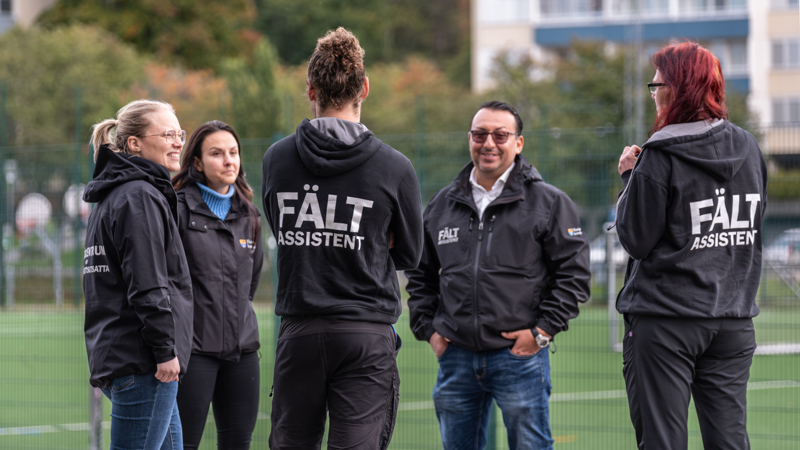 Bilden visar en grupp personer som står utomhus på en konstgräsplan med stängsel och flerbostadshus i bakgrunden. Personerna bär mörka jackor och tröjor med texten "FÄLT ASSISTENT" tryckt på ryggen och bröstet.