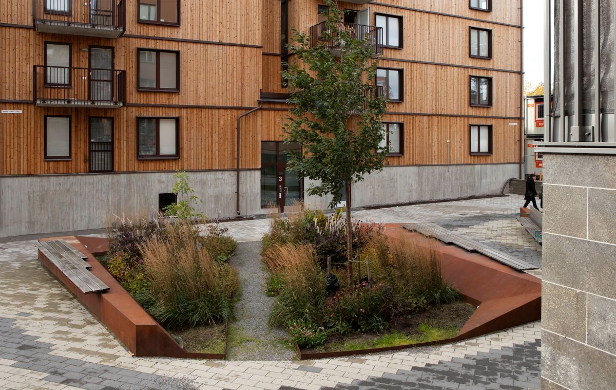 Växtlighet vid ett litet torg omgärdat av  gatustenar emd en mörk träfasad i bakgrunden