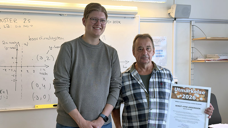 Två män i ett klassrum. Ena mannen håller i ett diplom.