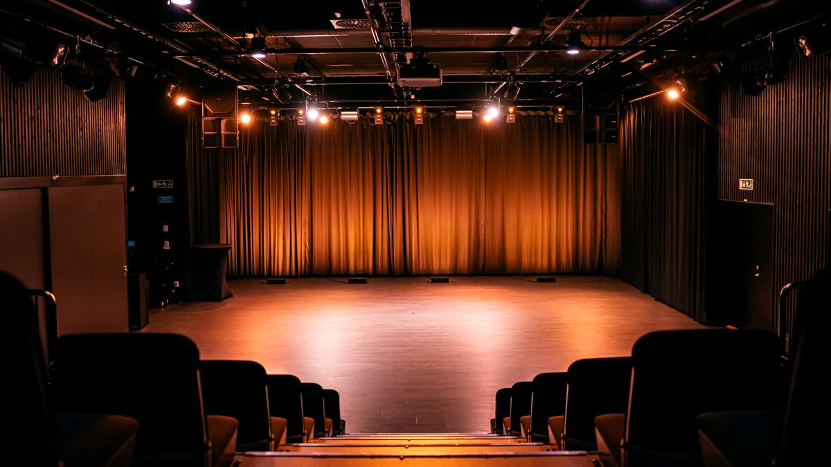 En tom scen med neddragna ridåer belyst av varma spotlights. Bilden är tagen från salongen där stolsraderna syns i förgrunden.